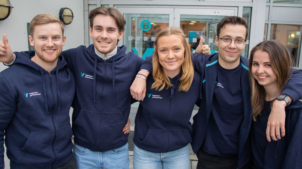 Fem glade studenter står arm i arm utenfor campus Stord. De har alle på seg blå hettejakke med høgskolen sin logo. De er studentambassadører og står klare til å ta imot nye studenter. 