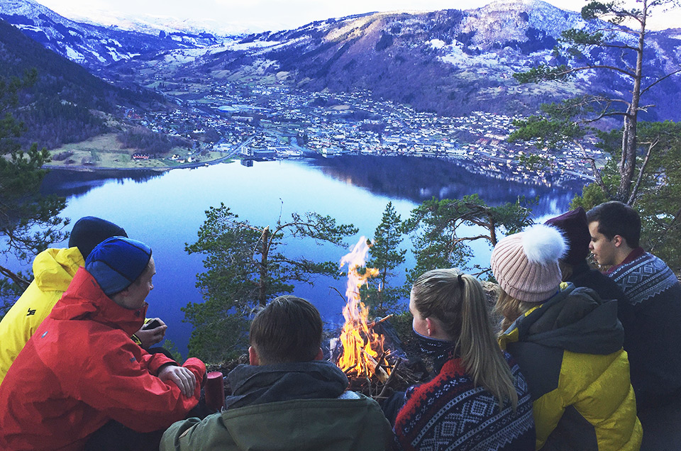 Studentar på båltur i skogen med fjorden og Sogndal i bakgrunnen. 